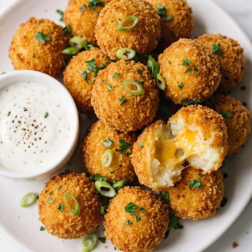 Golden panko-crusted mashed potato balls on a white plate with a small bowl of ranch dip