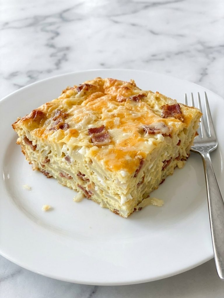 A white plate with a square of Amish breakfast casserole showing set egg, shredded hash browns, bacon and a golden cheese top