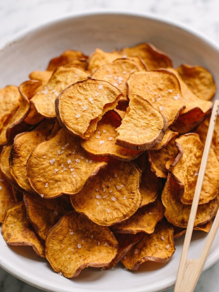 A white bowl piled high with crisp, golden baked sweet potato chips sprinkled with kosher salt