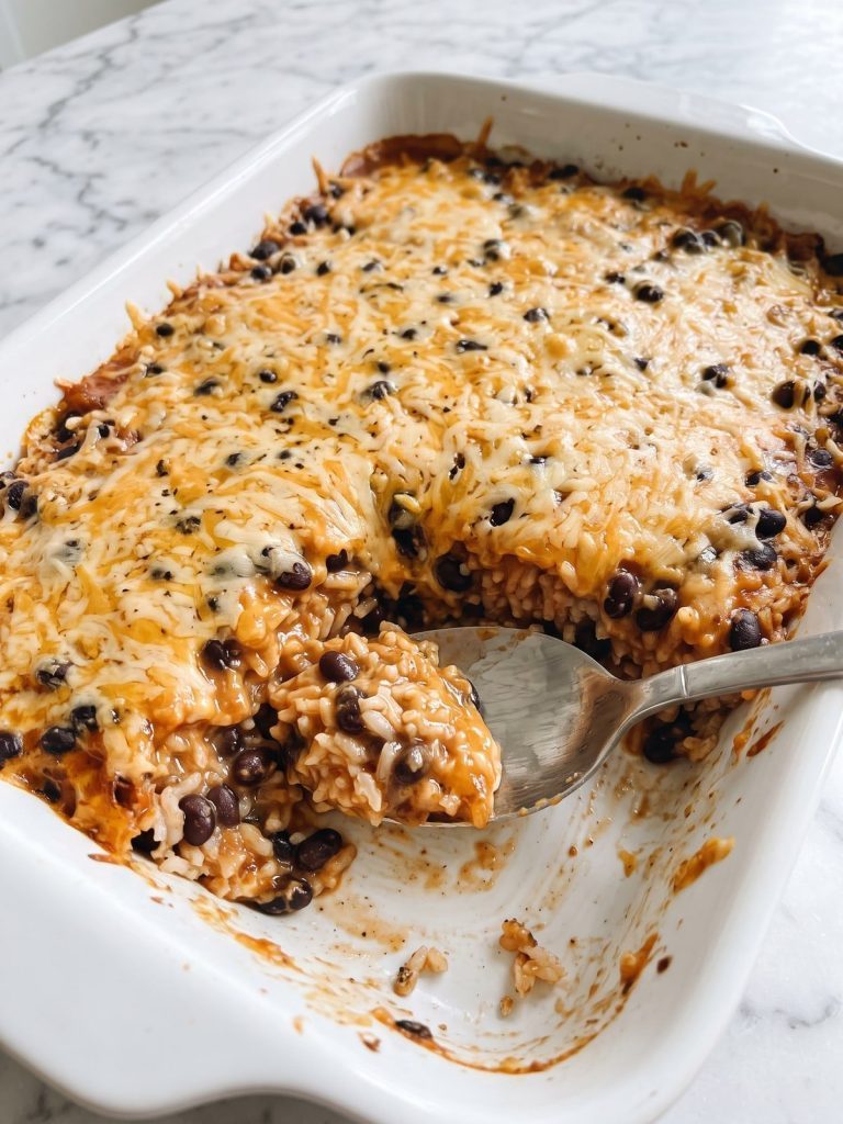 A baking dish of black bean rice casserole with melted cheddar on top, one serving scooped out to show black beans and rice in a red salsa sauce