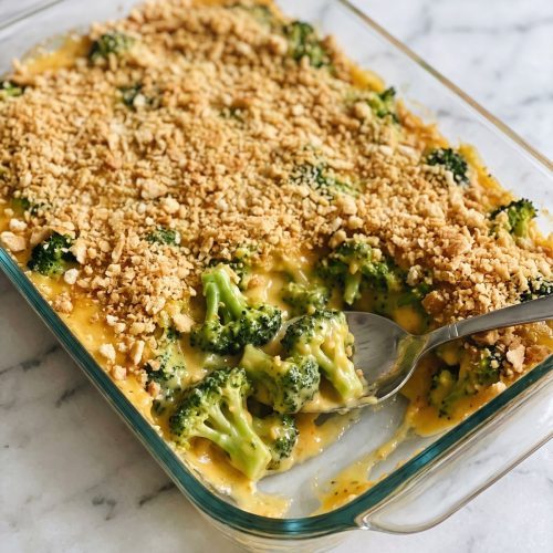 Broccoli casserole in a smooth yellow-orange cheese sauce under golden cracker crumbs in a glass 9x13 dish, one corner scooped out