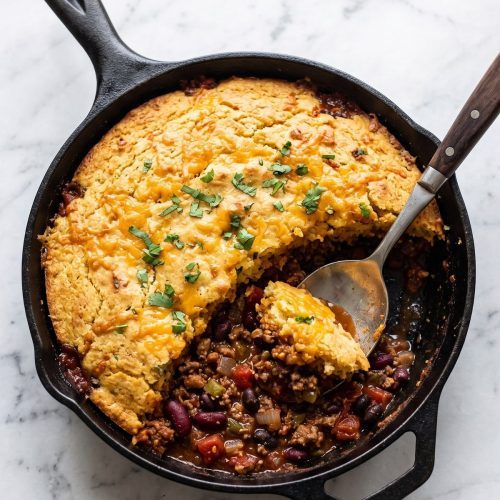 Chili cornbread casserole in a cast iron skillet with one serving scooped out to show the beef and bean chili