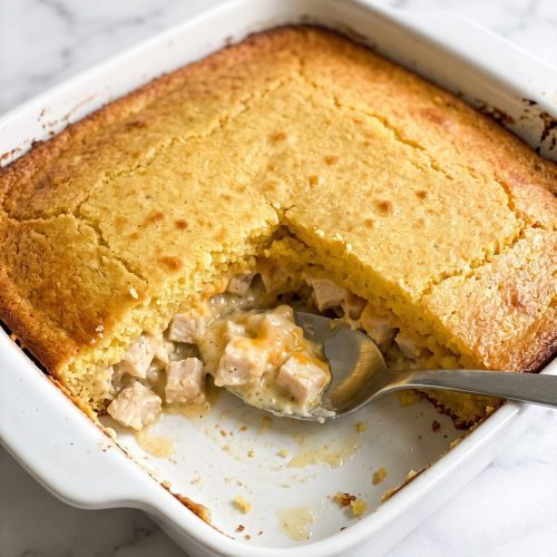 A baking dish of chicken cornbread casserole with one corner scooped out to show the creamy chicken filling under the golden cornbread