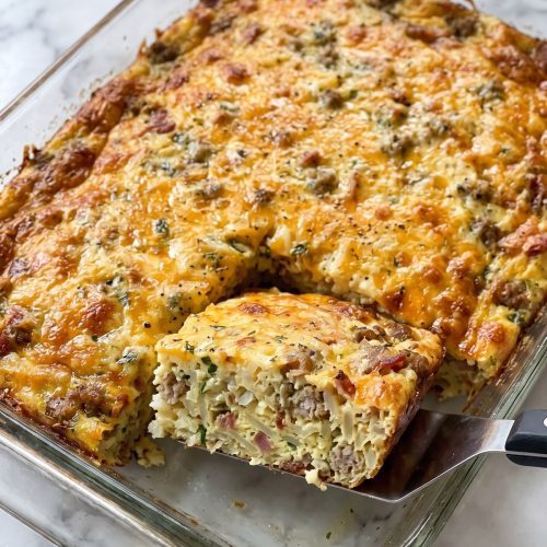 A crack breakfast casserole in a glass baking dish with one square lifted out, showing hash browns, sausage and bacon under browned cheddar