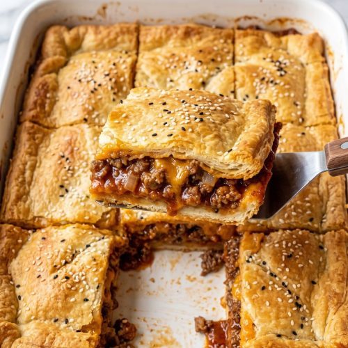 Crescent roll sloppy joe bake with a golden sesame-topped crust in a white baking dish, one square lifted to show the beef and cheddar filling