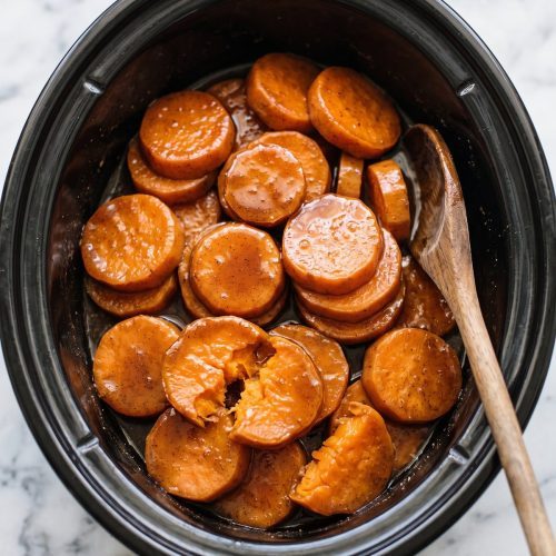Glossy candied sweet potato slices in brown sugar syrup in a black slow cooker with a wooden spoon