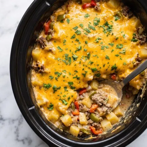 A black oval slow cooker of cheesesteak potato casserole with melted cheddar on top and one scoop taken out to show beef, peppers and potatoes