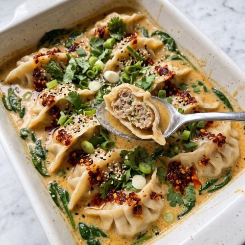 A white baking dish of baked dumplings in creamy coconut curry broth with spinach, cilantro, scallions and chili crisp