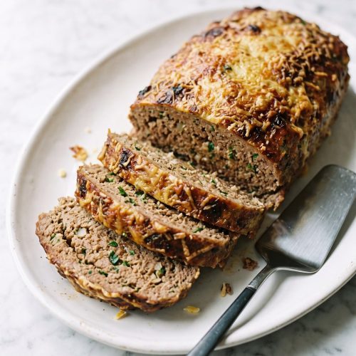 A whole garlic parmesan meatloaf on a white platter with a golden Parmesan top and three thick slices cut from the front