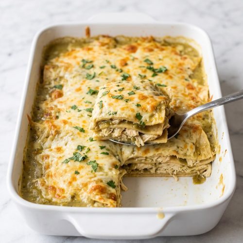 Baked green chili chicken enchilada casserole in a white dish with one serving scooped out to show the layers