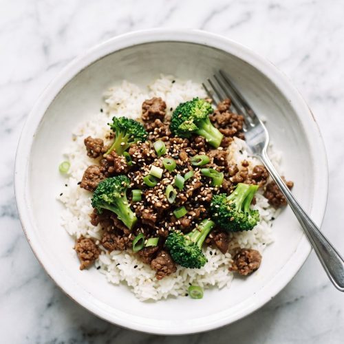 A bowl of white rice topped with ground beef and broccoli in a glossy soy sauce, with sesame seeds and green onions