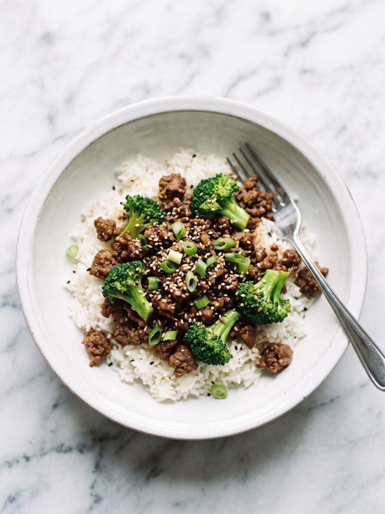 A bowl of white rice topped with ground beef and broccoli in a glossy soy sauce, with sesame seeds and green onions