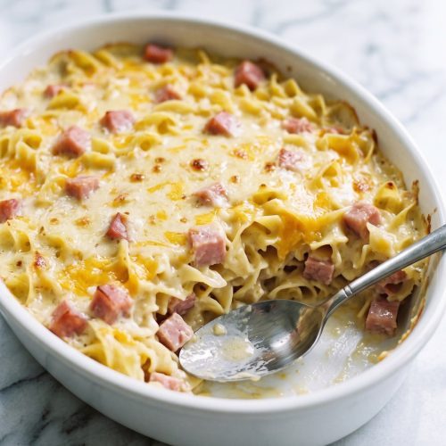 A white casserole dish of baked ham and noodle casserole with one serving scooped out to show the creamy egg noodle layers
