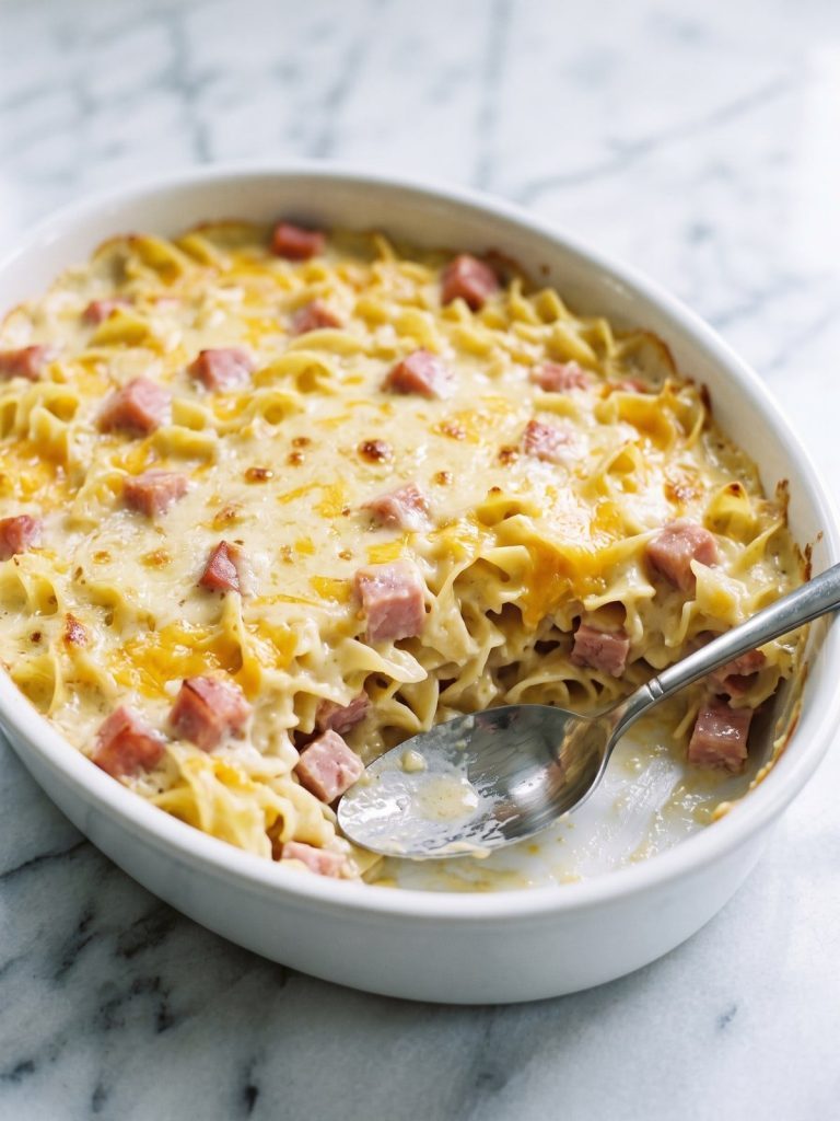 A white casserole dish of baked ham and noodle casserole with one serving scooped out to show the creamy egg noodle layers