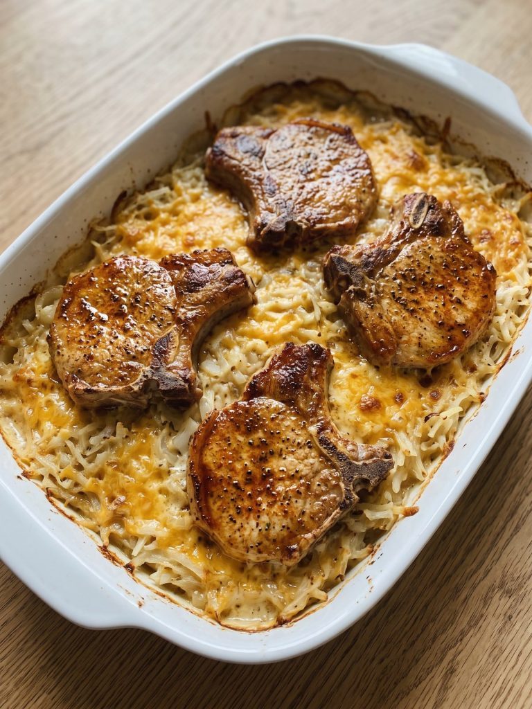 Four browned bone-in pork chops on a creamy, cheesy hash brown casserole in a baking dish