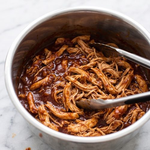 A stainless pressure cooker pot full of shredded chicken in glossy barbecue sauce with tongs