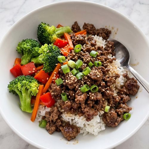 A bowl of Korean ground beef stir fry with broccoli, red bell pepper and carrots over white rice, topped with green onions and sesame seeds