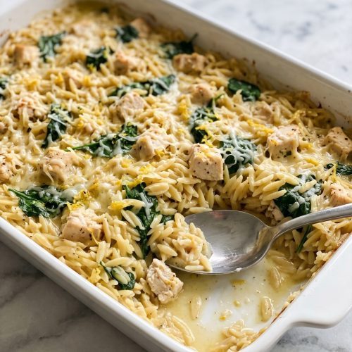 A white 9x13 baking dish of lemon chicken orzo bake with chicken, spinach and Parmesan, one serving scooped out
