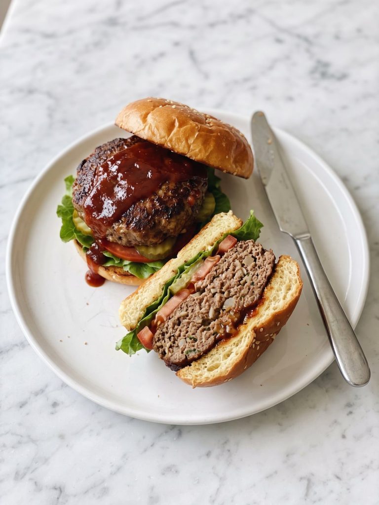 A glazed meatloaf burger on a bun with lettuce, tomato and pickles, cut in half on a white plate