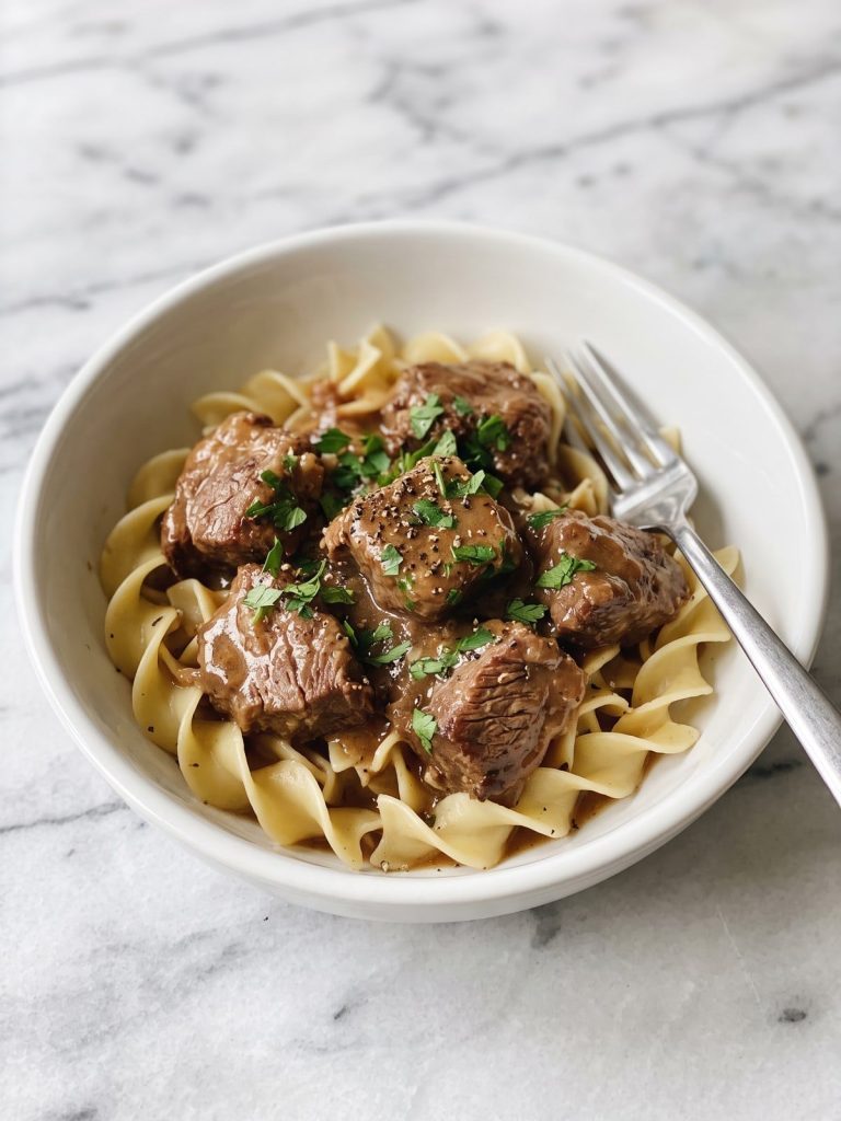 A bowl of wide egg noodles topped with tender chunks of beef in brown gravy and chopped parsley