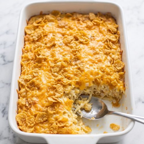 Cheesy party potatoes with a golden cornflake topping in a white baking dish with a serving spoon