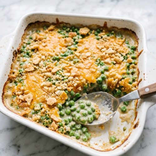 A square baking dish of green pea casserole with a golden cracker crumb topping, one corner scooped out to show the creamy peas