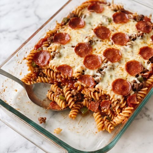 A glass baking dish of pizza pasta casserole with melted golden mozzarella and pepperoni slices, one serving scooped out to show rotini, beef and pepperoni in red sauce
