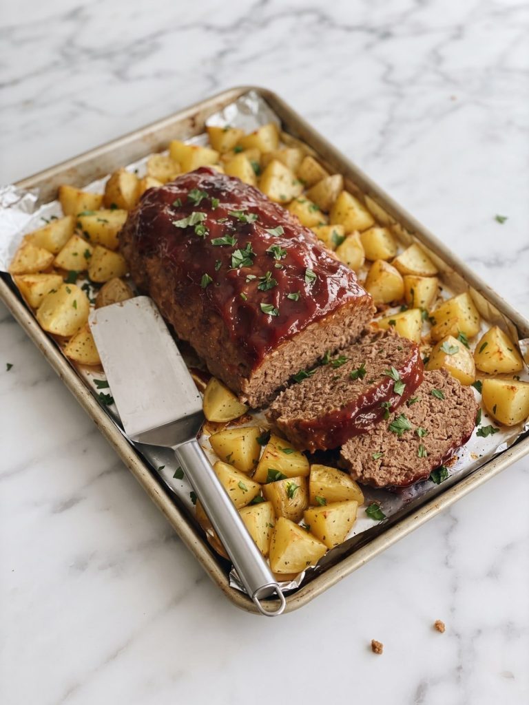 A glazed beef meatloaf with two slices cut, surrounded by golden roasted potatoes on a foil-lined sheet pan