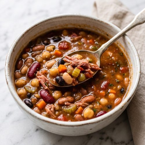 Bowl of slow cooker 15 bean soup with shredded ham, diced tomatoes and carrots, a spoon resting in the thick broth