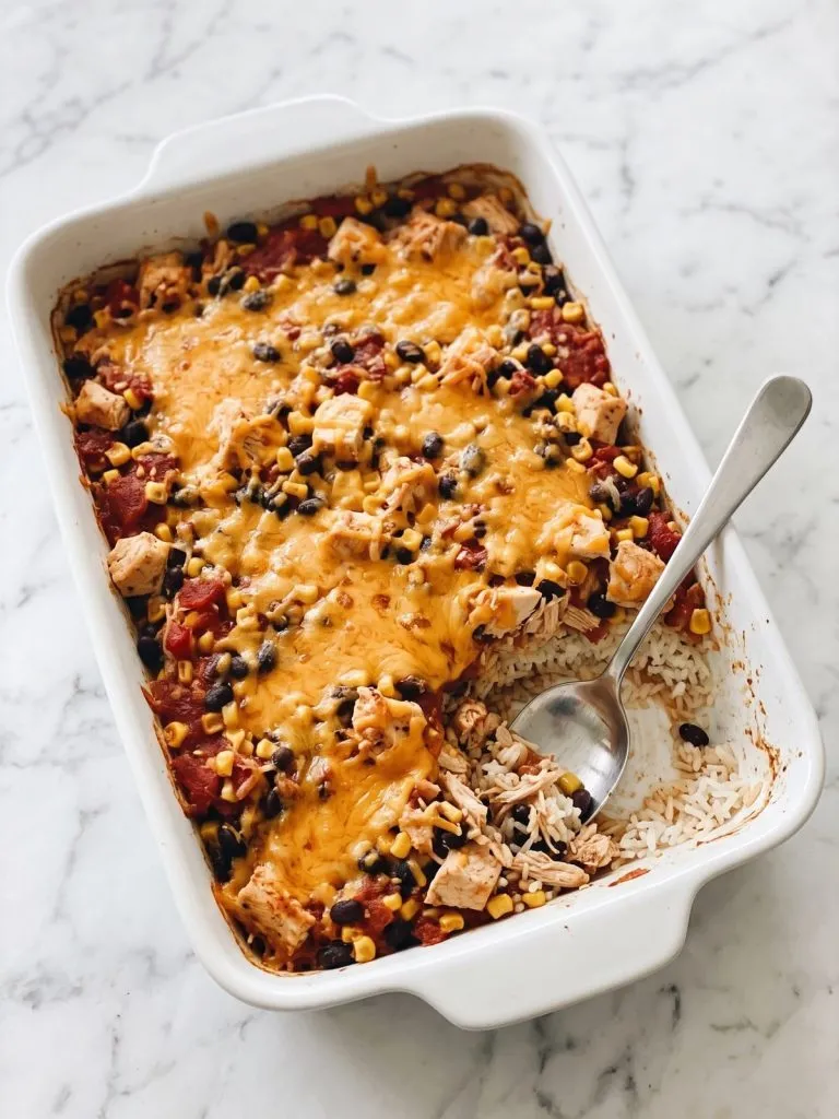 A baking dish of southwest chicken casserole with melted cheddar, black beans, corn and rice, one corner scooped out