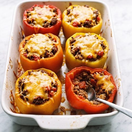 Six red, yellow and orange bell peppers stuffed with ground beef, rice and melted cheddar in a white baking dish