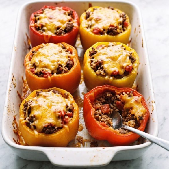 Six red, yellow and orange bell peppers stuffed with ground beef, rice and melted cheddar in a white baking dish