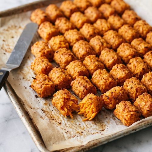Baked sweet potato tater tots on a lined sheet pan, one broken open to show the soft orange inside