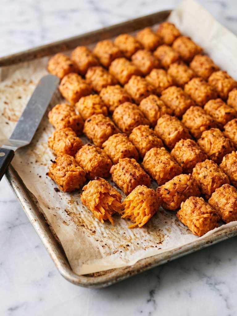 Baked sweet potato tater tots on a lined sheet pan, one broken open to show the soft orange inside