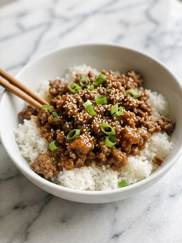 Close view of a white bowl of rice topped with glossy teriyaki ground beef, sesame seeds and green onions