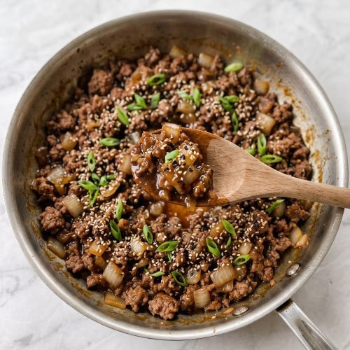 Teriyaki ground beef in a skillet with sesame seeds and green onions, with a spoon lifting a scoop