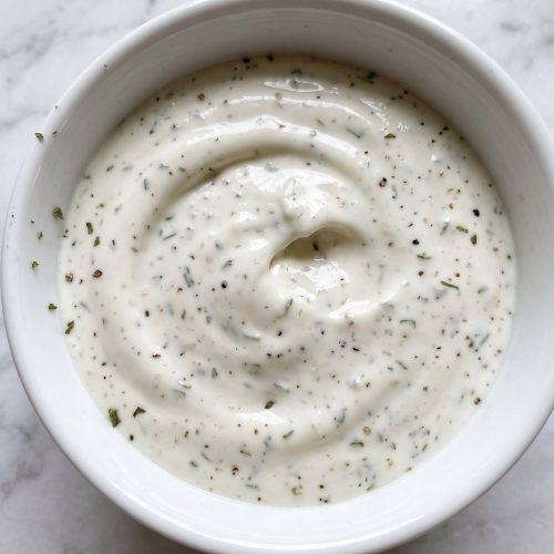 A white bowl of thin, creamy ranch dressing flecked with herbs
