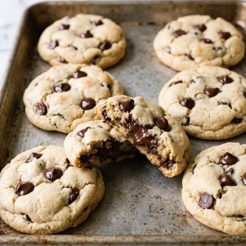 Thick, soft chocolate chip cookies on a baking sheet with one broken open