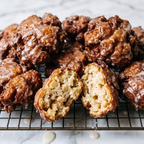 Glazed apple fritters on a wire rack with one broken open to show the apple pieces inside