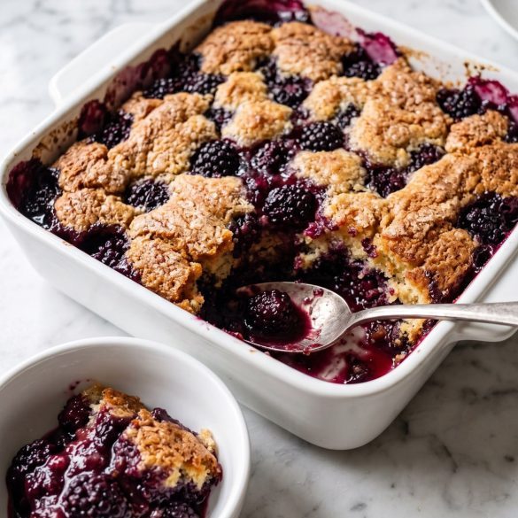 A baking dish of golden blackberry cobbler with dark berries and purple juices, one scoop served