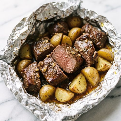 An opened foil pack with seared sirloin chunks and golden baby potatoes with herbs