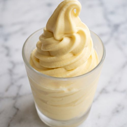 A tall swirl of pale yellow pineapple soft serve in a small frosted glass