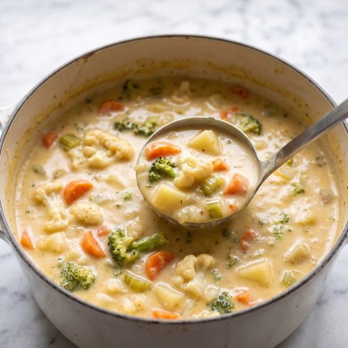 A pot of thick, creamy cheddar vegetable chowder with broccoli, potatoes and carrots, with a ladle